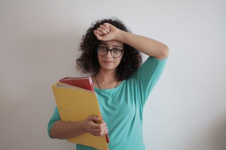 Accountant - Unhappy overworked female employee in glasses and casual clothes with folders of documents looking at camera with sadness and touching head while standing against white wall in contemporary office