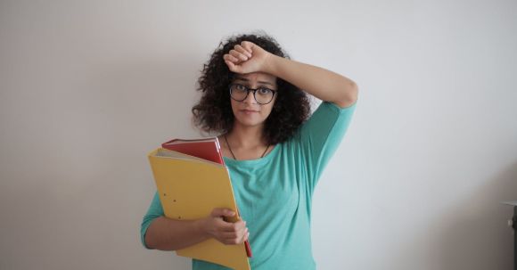 Accountant - Unhappy overworked female employee in glasses and casual clothes with folders of documents looking at camera with sadness and touching head while standing against white wall in contemporary office