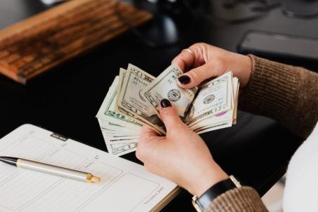 Accountant - Unrecognizable elegant female in sweater counting dollar bills while sitting at wooden table with planner and pen