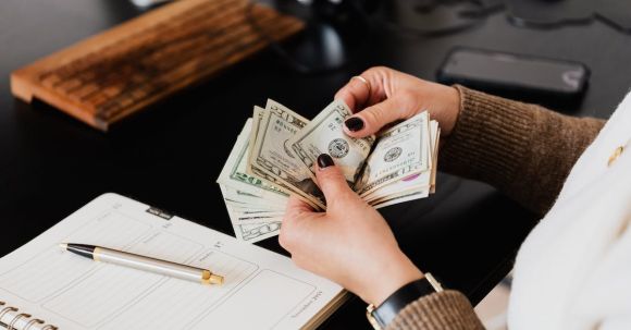 Accountant - Unrecognizable elegant female in sweater counting dollar bills while sitting at wooden table with planner and pen